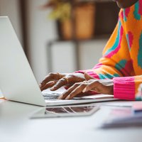Person wearing a colorful jumper sitting at a desk, focused on typing on a laptop. There is a mug, mobile phone and papers, visible on the desk.
