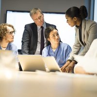 Group of healthcare professionals, including doctors and nurses, gathered around a desk. Some are working on laptops, while others are holding and reviewing documents in a collaborative discussion